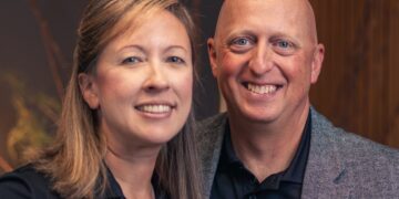 A professional portrait of a man and a woman standing close together and smiling. Both are wearing black polo shirts with a small white "A1" logo on the chest. The man, on the right, is bald and wearing a grey textured blazer over his polo. The woman, on the left, has shoulder-length light brown hair. They are posed against a warm, out-of-focus background that features dark wood paneling and decorative natural branches.