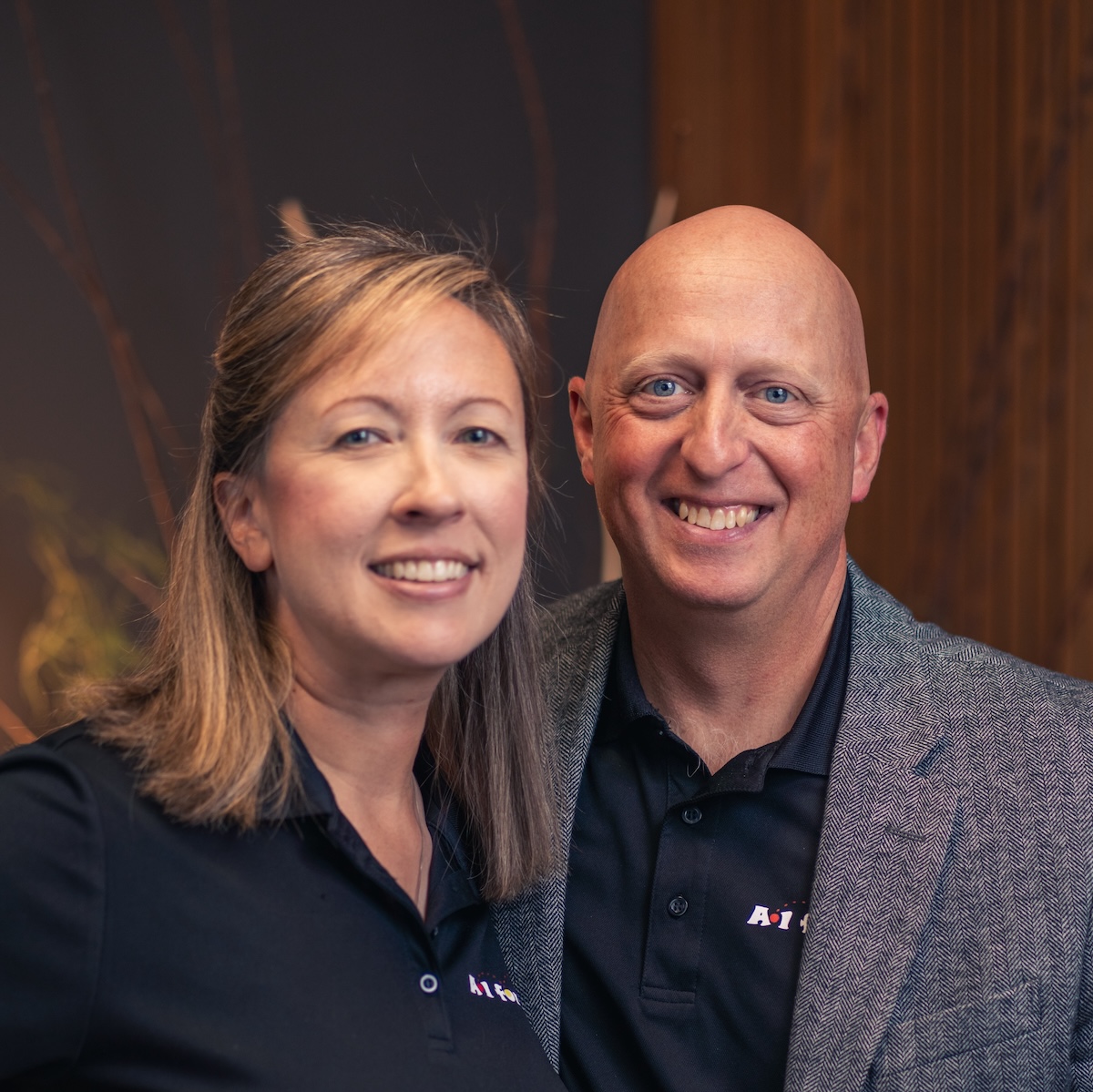 A professional portrait of a man and a woman standing close together and smiling. Both are wearing black polo shirts with a small white "A1" logo on the chest. The man, on the right, is bald and wearing a grey textured blazer over his polo. The woman, on the left, has shoulder-length light brown hair. They are posed against a warm, out-of-focus background that features dark wood paneling and decorative natural branches.