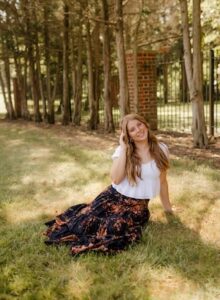 A young woman with long, reddish-blonde hair and a bright smile sits on a sun-dappled lawn. She is wearing a simple white short-sleeved top and a flowing, tiered maxi skirt with an orange and black floral or abstract print. She rests one hand lightly in her hair and the other on the grass beside her. The background is a soft-focus screen of slender trees, with a brick pillar and black wrought-iron gate visible behind them.