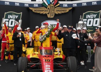 A celebratory group scene in Victory Circle at the Indianapolis Motor Speedway. In the center, Spanish driver Álex Palou, wearing a red and yellow racing suit and a matching cap, stands atop his red No. 10 Chip Ganassi Racing Honda. He is taking the traditional ceremonial swig of milk from a glass bottle while wearing a large, vibrant victory wreath of flowers. Surrounding him are jubilant team members and family, including team owner Chip Ganassi to his right. To the far right, the large, silver Borg-Warner Trophy is visible. The background features a large black display with "FOX SPORTS" and "INDY 500" logos.