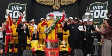 A celebratory group scene in Victory Circle at the Indianapolis Motor Speedway. In the center, Spanish driver Álex Palou, wearing a red and yellow racing suit and a matching cap, stands atop his red No. 10 Chip Ganassi Racing Honda. He is taking the traditional ceremonial swig of milk from a glass bottle while wearing a large, vibrant victory wreath of flowers. Surrounding him are jubilant team members and family, including team owner Chip Ganassi to his right. To the far right, the large, silver Borg-Warner Trophy is visible. The background features a large black display with "FOX SPORTS" and "INDY 500" logos.