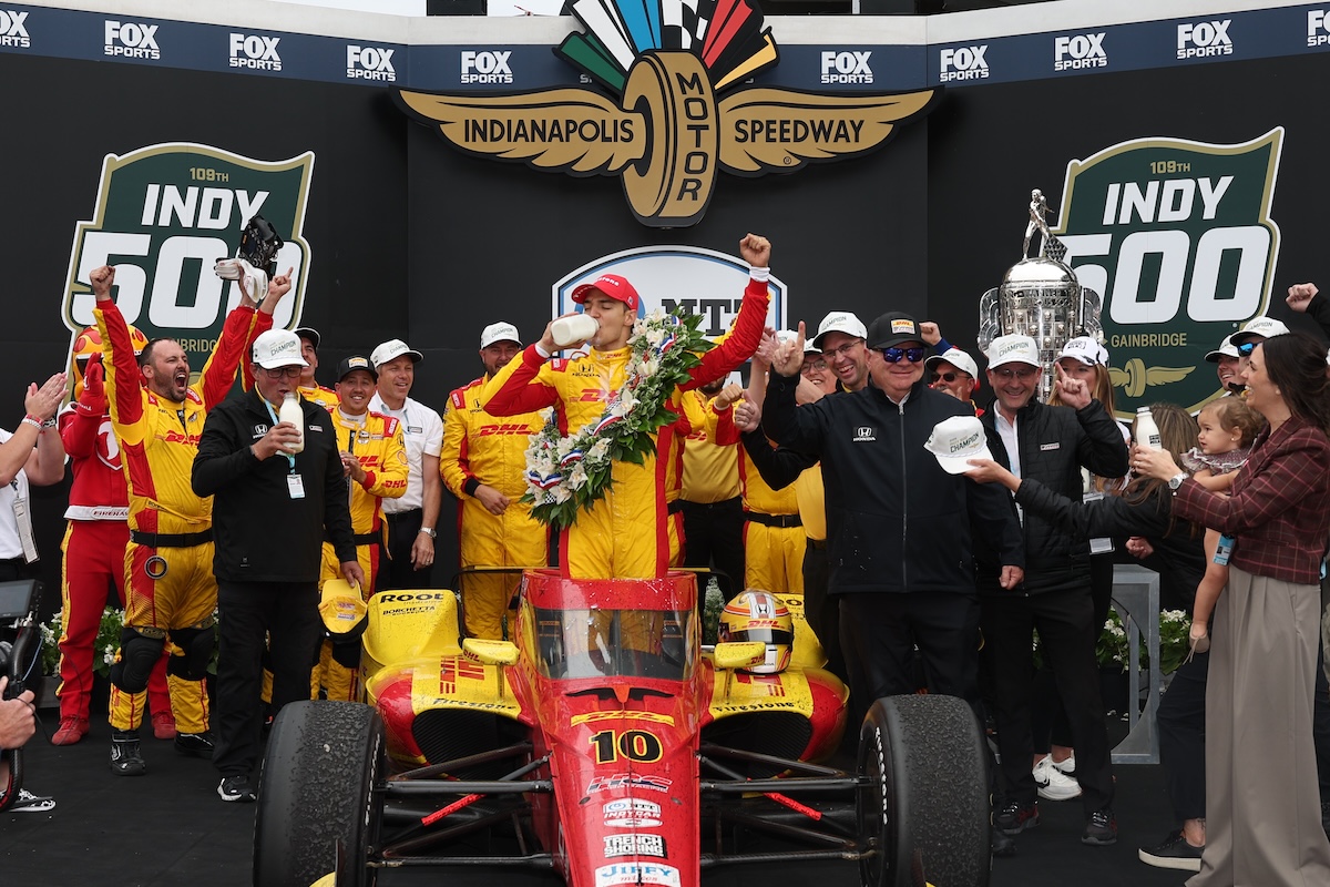 A celebratory group scene in Victory Circle at the Indianapolis Motor Speedway. In the center, Spanish driver Álex Palou, wearing a red and yellow racing suit and a matching cap, stands atop his red No. 10 Chip Ganassi Racing Honda. He is taking the traditional ceremonial swig of milk from a glass bottle while wearing a large, vibrant victory wreath of flowers. Surrounding him are jubilant team members and family, including team owner Chip Ganassi to his right. To the far right, the large, silver Borg-Warner Trophy is visible. The background features a large black display with "FOX SPORTS" and "INDY 500" logos.