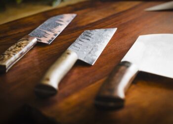 Three artisanal, hand-forged kitchen knives are showcased on a rich, dark-brown wooden cutting board. From left to right: a nakiri-style vegetable knife with a dark, textured blade and a marbled wood handle; a gyuto or chef’s knife with a hammered "tsuchi" finish and a light-colored wooden handle; and a large, wide-bladed cleaver with a polished finish and a dark, figured wood handle. The lighting is soft and dramatic, coming from the side and highlighting the unique textures and grain of both the metal and wood.