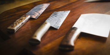 Three artisanal, hand-forged kitchen knives are showcased on a rich, dark-brown wooden cutting board. From left to right: a nakiri-style vegetable knife with a dark, textured blade and a marbled wood handle; a gyuto or chef’s knife with a hammered "tsuchi" finish and a light-colored wooden handle; and a large, wide-bladed cleaver with a polished finish and a dark, figured wood handle. The lighting is soft and dramatic, coming from the side and highlighting the unique textures and grain of both the metal and wood.