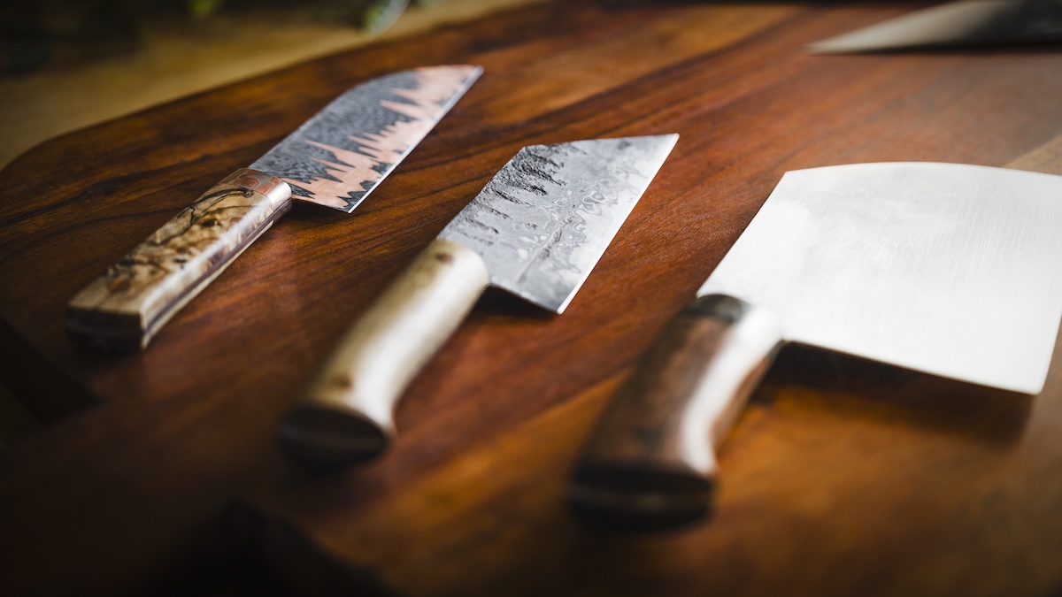 Three artisanal, hand-forged kitchen knives are showcased on a rich, dark-brown wooden cutting board. From left to right: a nakiri-style vegetable knife with a dark, textured blade and a marbled wood handle; a gyuto or chef’s knife with a hammered "tsuchi" finish and a light-colored wooden handle; and a large, wide-bladed cleaver with a polished finish and a dark, figured wood handle. The lighting is soft and dramatic, coming from the side and highlighting the unique textures and grain of both the metal and wood.