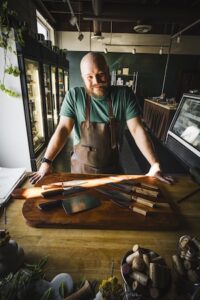A bearded man with a shaved head, wearing a green t-shirt and a heavy-duty brown leather apron, stands behind a wooden counter. He leans forward with his hands on the counter, smiling warmly at the camera. In front of him, a collection of artisanal kitchen knives with various blade shapes and wooden handles is displayed on a large, dark wood cutting board. The background shows the interior of a shop or workshop with refrigerators and display cases.