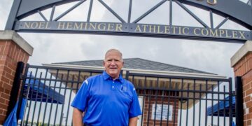 A man in a blue polo shirt stands smiling in front of a black gated entrance. Above him is a large, black metal archway with gold lettering that reads "GRIZZLY FIELDS" and a smaller sign below it for the "NOEL HEMINGER ATHLETIC COMPLEX." Behind the gates, a brick building and athletic field equipment are visible under a cloudy sky.