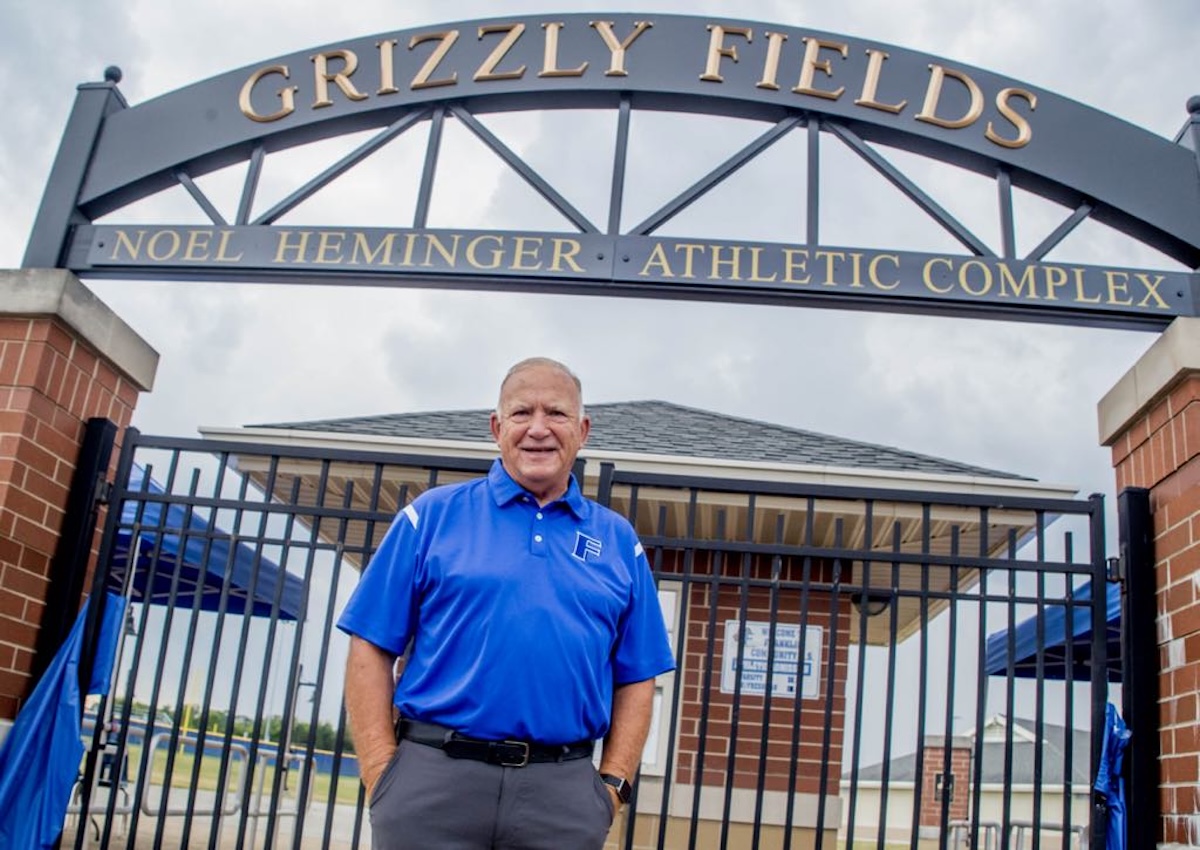 A man in a blue polo shirt stands smiling in front of a black gated entrance. Above him is a large, black metal archway with gold lettering that reads "GRIZZLY FIELDS" and a smaller sign below it for the "NOEL HEMINGER ATHLETIC COMPLEX." Behind the gates, a brick building and athletic field equipment are visible under a cloudy sky.