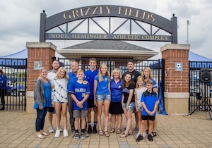 A group of twelve people, including adults and children of various ages, pose together in front of a gated entrance. Most are wearing blue apparel with local school logos. They are positioned beneath a large black metal arch that reads "GRIZZLY FIELDS" and "NOEL HEMINGER ATHLETIC COMPLEX." The group is standing on a paved area with brick pillars on either side of the black iron gate.