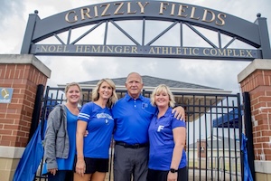 Four individuals—three women and one man—stand together in a close group, smiling for the camera. The man stands in the center with his arms around two of the women. They are all dressed in blue apparel, some featuring the "F" logo for Franklin Community High School. They are positioned directly under the black metal archway of the Noel Heminger Athletic Complex at Grizzly Fields. The background shows the black iron gates and brick pillars of the entrance under an overcast sky.