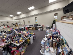 A high-angle, wide shot of an office space transformed into a massive toy collection center. Multiple long folding tables are overflowing with hundreds of new, boxed toys, including action figures, board games, and craft kits. In the background, additional tables hold more donations against a neutral office wall with high windows. The sheer volume of toys fills a significant portion of the room, leaving only narrow walkways.