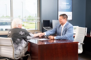 A smiling man in a blue patterned blazer, identified as Bob Haymond, sits behind a dark wood desk and shakes hands with an older woman seated across from him. The woman, seen from behind, has white hair and wears a grey and white floral top. The office is modern and bright, with large windows showing an outdoor parking area, a dark navy blue accent wall, and professional equipment like a laptop and monitor on the desk.