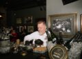 A medium shot of a chef from Huse Culinary, wearing black gloves and a white uniform, meticulously preparing appetizers at a black-clothed bar. He is placing small, upright pastry shells onto a metal rack, ready to be filled. In the foreground, a luxurious spread is showcased: a gold-rimmed tin of Imperial Caviar Osietra sits on ice next to chilled bottles of JCB No. 21 Champagne and JCB No. 69 Rosé. The setting is a moody, vintage-inspired speakeasy, likely the 1933 Lounge in Indianapolis. The walls are decorated with framed black-and-white historical photos of crowded bars and a "PROHIBITION ENDS AT LAST" tin sign, enhancing the authentic Repeal Day atmosphere.