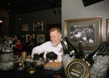 A medium shot of a chef from Huse Culinary, wearing black gloves and a white uniform, meticulously preparing appetizers at a black-clothed bar. He is placing small, upright pastry shells onto a metal rack, ready to be filled. In the foreground, a luxurious spread is showcased: a gold-rimmed tin of Imperial Caviar Osietra sits on ice next to chilled bottles of JCB No. 21 Champagne and JCB No. 69 Rosé. The setting is a moody, vintage-inspired speakeasy, likely the 1933 Lounge in Indianapolis. The walls are decorated with framed black-and-white historical photos of crowded bars and a "PROHIBITION ENDS AT LAST" tin sign, enhancing the authentic Repeal Day atmosphere.