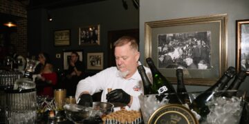 A medium shot of a chef from Huse Culinary, wearing black gloves and a white uniform, meticulously preparing appetizers at a black-clothed bar. He is placing small, upright pastry shells onto a metal rack, ready to be filled. In the foreground, a luxurious spread is showcased: a gold-rimmed tin of Imperial Caviar Osietra sits on ice next to chilled bottles of JCB No. 21 Champagne and JCB No. 69 Rosé. The setting is a moody, vintage-inspired speakeasy, likely the 1933 Lounge in Indianapolis. The walls are decorated with framed black-and-white historical photos of crowded bars and a "PROHIBITION ENDS AT LAST" tin sign, enhancing the authentic Repeal Day atmosphere.