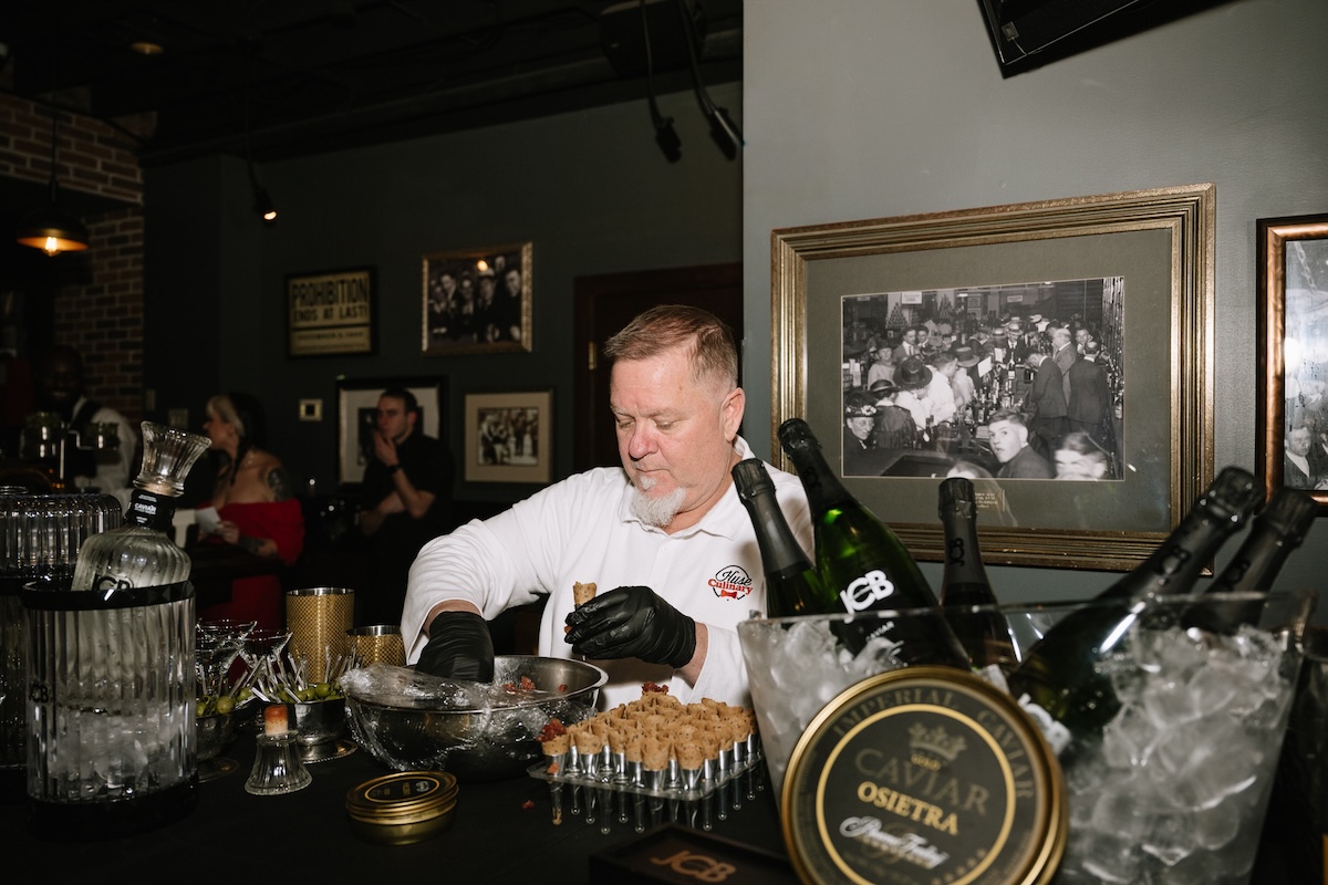 A medium shot of a chef from Huse Culinary, wearing black gloves and a white uniform, meticulously preparing appetizers at a black-clothed bar. He is placing small, upright pastry shells onto a metal rack, ready to be filled. In the foreground, a luxurious spread is showcased: a gold-rimmed tin of Imperial Caviar Osietra sits on ice next to chilled bottles of JCB No. 21 Champagne and JCB No. 69 Rosé. The setting is a moody, vintage-inspired speakeasy, likely the 1933 Lounge in Indianapolis. The walls are decorated with framed black-and-white historical photos of crowded bars and a "PROHIBITION ENDS AT LAST" tin sign, enhancing the authentic Repeal Day atmosphere.