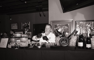 A dramatic black-and-white photo captures Michael Christensen, Director of Culinary Operations for Huse Culinary, meticulously plating appetizers at a vintage-inspired speakeasy event. Christensen, wearing a white chef's jacket and black gloves, is focused on placing small pastry shells onto a tiered metal serving rack. The bar in front of him features a luxurious spread: a large, branded tin of Imperial Caviar Osietra sits on a bed of ice, flanked by chilled bottles of JCB No. 21 Champagne and JCB No. 69 Rosé. The background is adorned with framed historical photographs and a "PROHIBITION ENDS AT LAST" sign, evoking the classic atmosphere of a Repeal Day celebration at the 1933 Lounge.