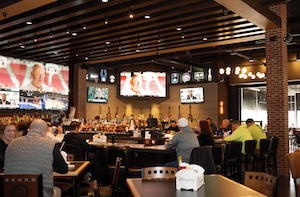 The interior of a spacious, modern sports bar with high dark ceilings and industrial-style wooden beams. Multiple massive flat-screen televisions are mounted across the walls, displaying various sports games. Patrons are seated at a long, dark-top bar and surrounding high-top tables. 