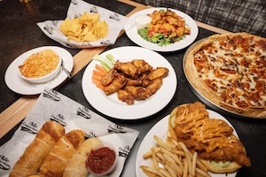 A top-down view of a dark wooden table covered with several plates of popular bar food. The spread includes a traditional pepperoni and sausage pizza, a plate of Hermanaki® wings with celery and carrots, a basket of stuffed breadsticks with a side of marinara, a bowl of creamy buffalo chicken dip with tortilla chips, and a fried fish sandwich served with a side of golden French fries.