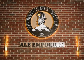 The Ale Emporium logo is prominently displayed on a red brick wall, lit by warm, vertical light fixtures on either side. The circular logo features a profile of a man in a helmet, with the French motto "LE BON TEMPS VIENDRA" (The good times will come) arching above him. Below the circular emblem, the words "ALE EMPORIUM" are cut out in large, backlit metal letters against the brick.
