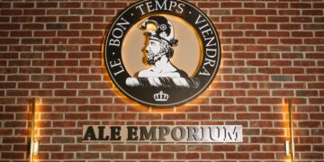 The Ale Emporium logo is prominently displayed on a red brick wall, lit by warm, vertical light fixtures on either side. The circular logo features a profile of a man in a helmet, with the French motto "LE BON TEMPS VIENDRA" (The good times will come) arching above him. Below the circular emblem, the words "ALE EMPORIUM" are cut out in large, backlit metal letters against the brick.
