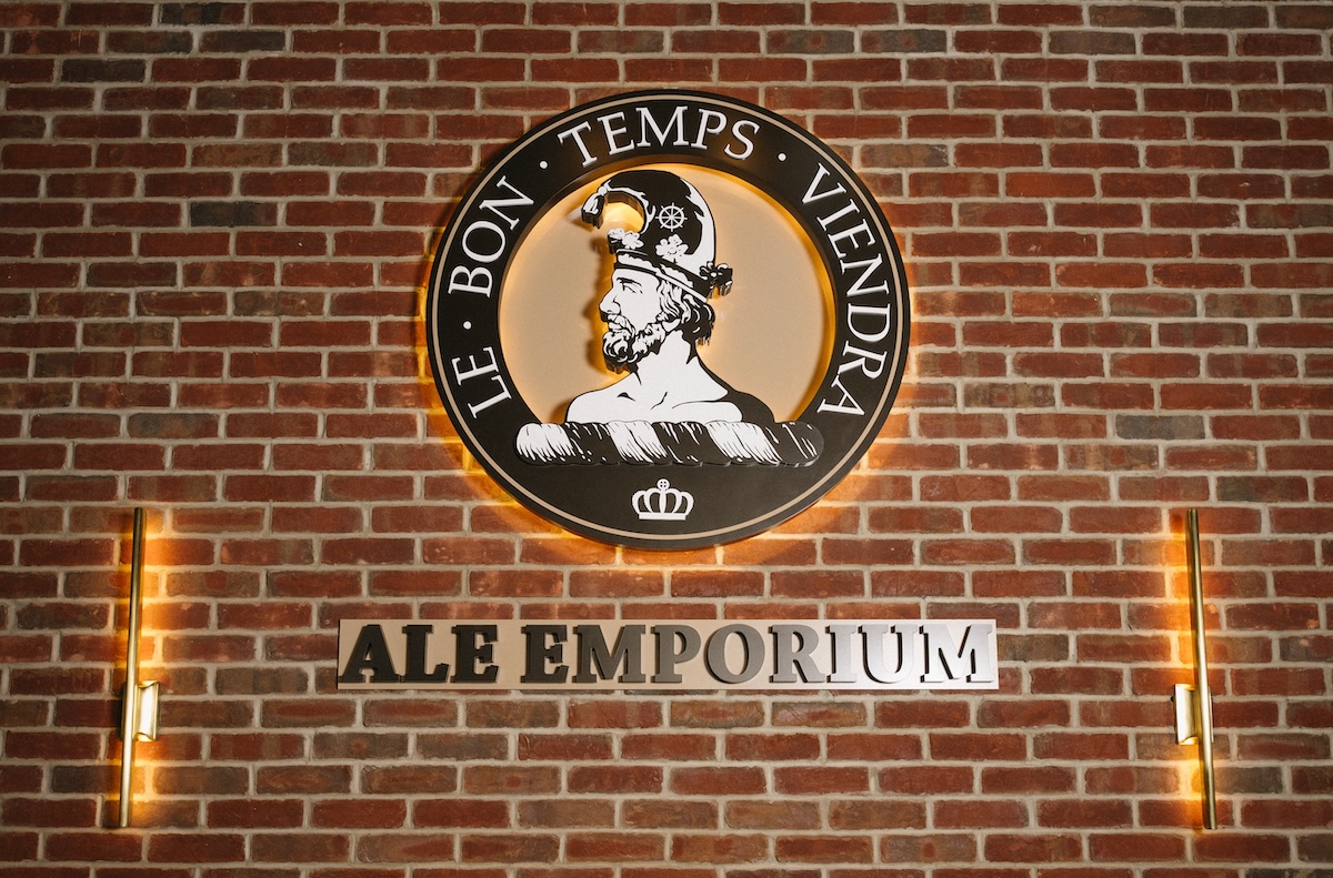 The Ale Emporium logo is prominently displayed on a red brick wall, lit by warm, vertical light fixtures on either side. The circular logo features a profile of a man in a helmet, with the French motto "LE BON TEMPS VIENDRA" (The good times will come) arching above him. Below the circular emblem, the words "ALE EMPORIUM" are cut out in large, backlit metal letters against the brick.