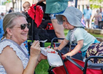 A woman with blonde hair pulled back in a ponytail and wearing sunglasses smiles warmly while holding a white foam takeout container filled with strawberries, ice cream, and whipped topping. She is sitting outdoors on a grassy area next to a young child in a red wagon. The child, wearing a light blue t-shirt and a grey sun hat, leans forward over the edge of the wagon, looking intently at the food. Other people and a red stroller are visible in the blurred background.
