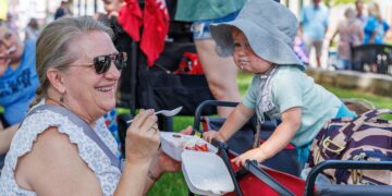A woman with blonde hair pulled back in a ponytail and wearing sunglasses smiles warmly while holding a white foam takeout container filled with strawberries, ice cream, and whipped topping. She is sitting outdoors on a grassy area next to a young child in a red wagon. The child, wearing a light blue t-shirt and a grey sun hat, leans forward over the edge of the wagon, looking intently at the food. Other people and a red stroller are visible in the blurred background.