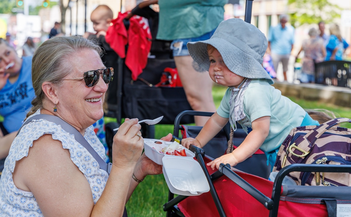 A woman with blonde hair pulled back in a ponytail and wearing sunglasses smiles warmly while holding a white foam takeout container filled with strawberries, ice cream, and whipped topping. She is sitting outdoors on a grassy area next to a young child in a red wagon. The child, wearing a light blue t-shirt and a grey sun hat, leans forward over the edge of the wagon, looking intently at the food. Other people and a red stroller are visible in the blurred background.