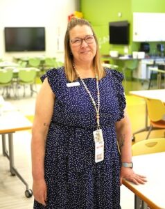 A woman with light brown hair and glasses smiles for a portrait. She is wearing a navy blue dress with a white speckled pattern, a white name tag, and a colorful beaded lanyard with a photo ID. She is standing in a modern, brightly lit classroom or learning center, with green and yellow accents and several desks and chairs visible in the background.