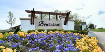 A vibrant, eye-level shot of the entrance to the Bonterra community by M/I Homes. A large, modern sign with "Bonterra" in black script is mounted on a light-colored stone wall, topped with a dark wood pergola. In the foreground, a dense field of bright yellow and purple pansies fills the frame, leading the eye toward the community entrance. An M/I Homes logo is visible on a smaller sign in the background, set against a bright, cloudy sky and lush green trees.