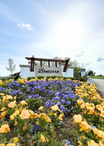 A vibrant, eye-level shot of the entrance to the Bonterra community by M/I Homes. A large, modern sign with "Bonterra" in black script is mounted on a light-colored stone wall, topped with a dark wood pergola. In the foreground, a dense field of bright yellow and purple pansies fills the frame, leading the eye toward the community entrance. An M/I Homes logo is visible on a smaller sign in the background, set against a bright, cloudy sky and lush green trees.