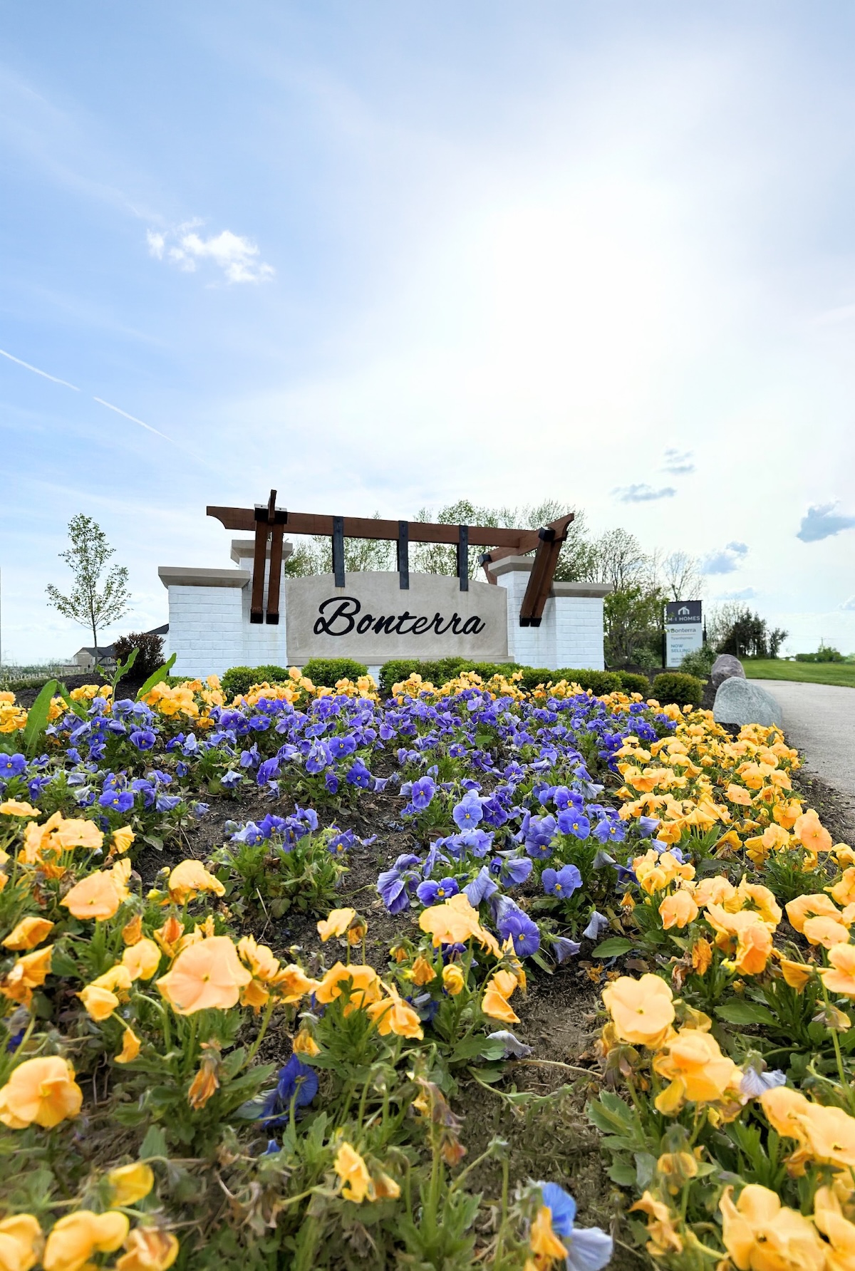 A vibrant, eye-level shot of the entrance to the Bonterra community by M/I Homes. A large, modern sign with "Bonterra" in black script is mounted on a light-colored stone wall, topped with a dark wood pergola. In the foreground, a dense field of bright yellow and purple pansies fills the frame, leading the eye toward the community entrance. An M/I Homes logo is visible on a smaller sign in the background, set against a bright, cloudy sky and lush green trees.