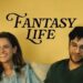 Amanda Peet (left) and Matthew Shear (right) sit on a couch in front of a yellow wall. Peet, with long brown hair and a denim shirt, laughs while looking off-camera. Shear wears glasses and a dark green shirt, looking down thoughtfully. The words "FANTASY LIFE" are printed in a stylized black font on the wall between them.