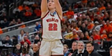 A female basketball player in a white Lawrence North Wildcats jersey with the number 22 in red is captured mid-shot on a basketball court. She is looking up at the hoop with both arms raised in a follow-through motion. She wears white shorts with green and red trim. In the background, a large, blurred crowd of spectators fills the stadium seats, many wearing orange, while several men in black athletic gear sit on the sidelines.