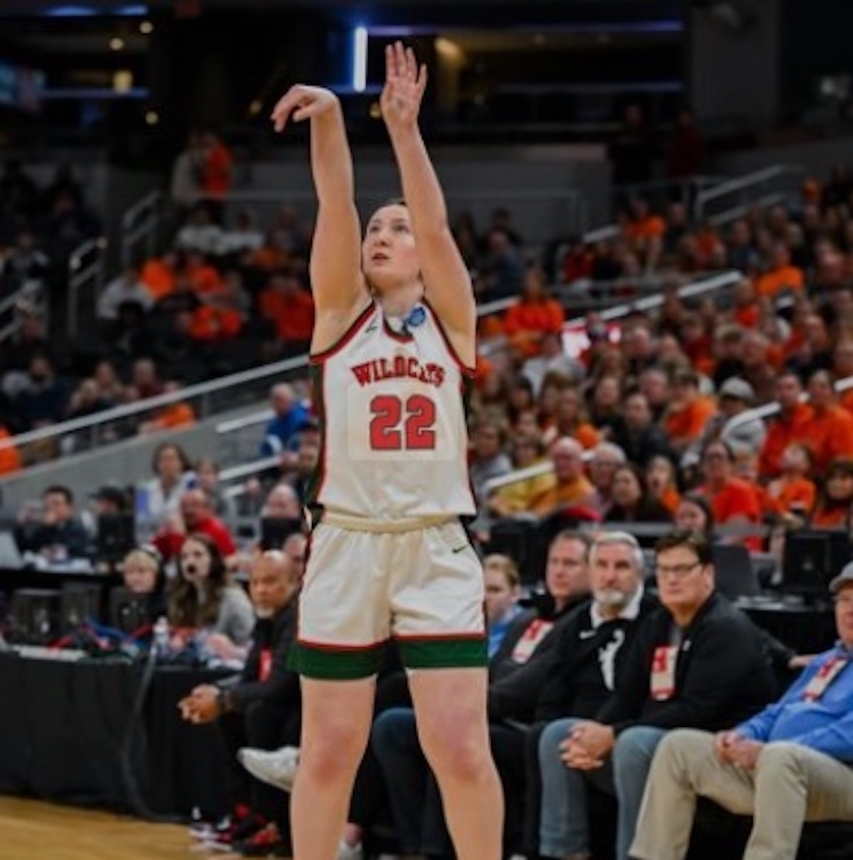 A female basketball player in a white Lawrence North Wildcats jersey with the number 22 in red is captured mid-shot on a basketball court. She is looking up at the hoop with both arms raised in a follow-through motion. She wears white shorts with green and red trim. In the background, a large, blurred crowd of spectators fills the stadium seats, many wearing orange, while several men in black athletic gear sit on the sidelines.