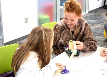 Two young girls are seated at a white table in a brightly lit, modern classroom or activity center. The girl in the foreground has long, straight brown hair and is looking towards her companion. The girl on the right, with reddish hair pulled into a messy bun, wears a camouflage-patterned hoodie and is laughing joyfully. She is holding a small, handmade toy car constructed from recycled materials like cardboard, plastic wheels, and tape. A pair of blue scissors and other craft supplies are scattered on the table.