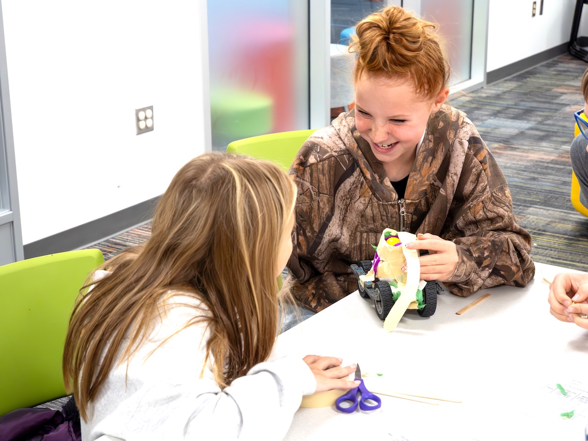 Two young girls are seated at a white table in a brightly lit, modern classroom or activity center. The girl in the foreground has long, straight brown hair and is looking towards her companion. The girl on the right, with reddish hair pulled into a messy bun, wears a camouflage-patterned hoodie and is laughing joyfully. She is holding a small, handmade toy car constructed from recycled materials like cardboard, plastic wheels, and tape. A pair of blue scissors and other craft supplies are scattered on the table.