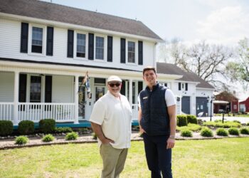 Two men, one older in a tan shirt and cap and one younger in a navy vest, stand together on a green lawn in front of a two-story white farmhouse with black shutters and a wrap-around porch.