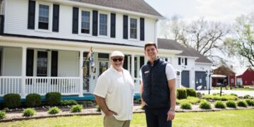 Two men, one older in a tan shirt and cap and one younger in a navy vest, stand together on a green lawn in front of a two-story white farmhouse with black shutters and a wrap-around porch.