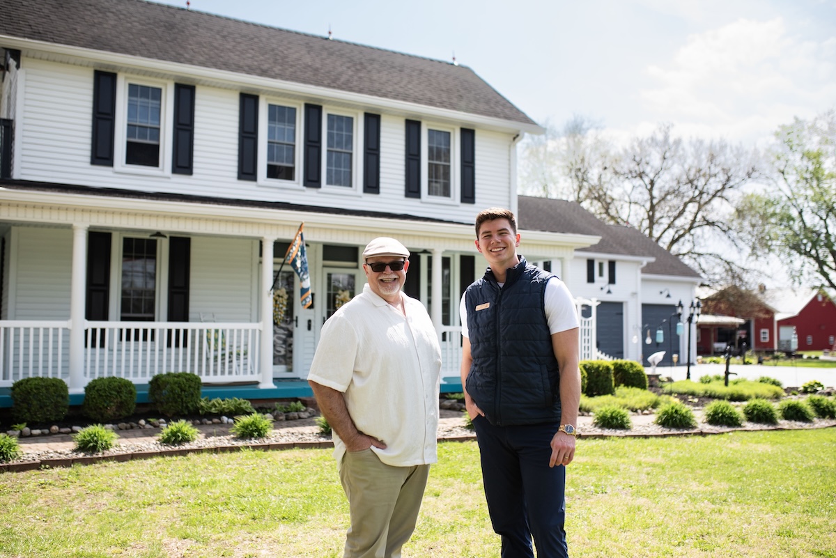 Two men, one older in a tan shirt and cap and one younger in a navy vest, stand together on a green lawn in front of a two-story white farmhouse with black shutters and a wrap-around porch.