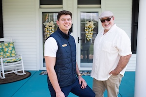 A younger man in a navy puffer vest and an older man in a cream shirt and flat cap stand together on a bright blue porch. Behind them are white double doors decorated with yellow floral wreaths and a white rocking chair with a patterned cushion. Both men are smiling at the camera.