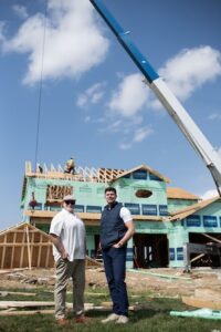 An older man in a flat cap and a younger man in a navy puffer vest stand side-by-side on a construction site. Behind them, a two-story house is under construction, wrapped in green weather barrier with exposed wood framing. A large blue and white crane boom extends diagonally across the bright blue sky, and a worker is visible on the roof.