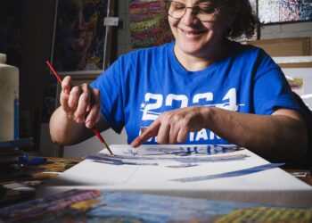 A smiling woman with curly hair and glasses, wearing a royal blue t-shirt with "2024" printed on it, is deeply focused on her artwork. She is seated at a table, using a paintbrush with her right hand and pointing to a specific detail on a large white canvas with her left. The canvas is a work-in-progress mixed media piece with various blue paper cutouts and sketches. The background wall is covered with colorful, textured paintings and posters, including a prominent portrait of John Lennon. In the foreground, another vibrant, multi-colored painting is visible, out of focus.