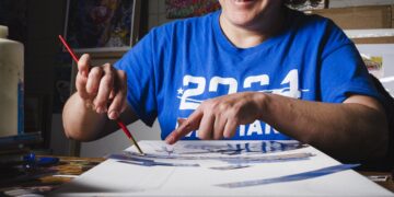 A smiling woman with curly hair and glasses, wearing a royal blue t-shirt with "2024" printed on it, is deeply focused on her artwork. She is seated at a table, using a paintbrush with her right hand and pointing to a specific detail on a large white canvas with her left. The canvas is a work-in-progress mixed media piece with various blue paper cutouts and sketches. The background wall is covered with colorful, textured paintings and posters, including a prominent portrait of John Lennon. In the foreground, another vibrant, multi-colored painting is visible, out of focus.