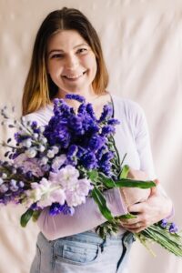 A smiling woman with long brown hair stands against a neutral, draped fabric backdrop. She is wearing a light purple long-sleeved shirt and blue jeans, holding a large, lush bouquet of purple and lilac flowers close to her chest.
