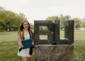 A medium full-shot of a smiling young woman celebrating her graduation at Butler University. She stands on a green lawn, wearing a short white dress and a black graduation stole with gold and blue trim. She holds a navy blue Butler University diploma cover. Next to her is the iconic BU statue, a large metallic sculpture of the university's initials resting on a massive granite base. Lush green trees and campus buildings are visible in the soft-focus background under a bright, overcast sky.