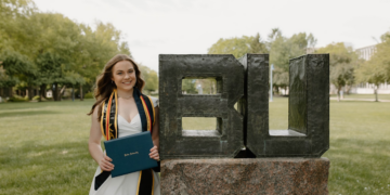 A medium full-shot of a smiling young woman celebrating her graduation at Butler University. She stands on a green lawn, wearing a short white dress and a black graduation stole with gold and blue trim. She holds a navy blue Butler University diploma cover. Next to her is the iconic BU statue, a large metallic sculpture of the university's initials resting on a massive granite base. Lush green trees and campus buildings are visible in the soft-focus background under a bright, overcast sky.