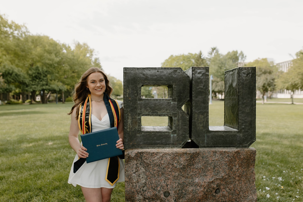 A medium full-shot of a smiling young woman celebrating her graduation at Butler University. She stands on a green lawn, wearing a short white dress and a black graduation stole with gold and blue trim. She holds a navy blue Butler University diploma cover. Next to her is the iconic BU statue, a large metallic sculpture of the university's initials resting on a massive granite base. Lush green trees and campus buildings are visible in the soft-focus background under a bright, overcast sky.