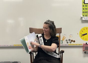A smiling young woman, serving as a 500 Festival Princess for 2026, sits in a wooden chair in a classroom. She is wearing a black dress, a sparkling tiara, and her official white sash as she reads a colorful children's picture book aloud. The background features typical classroom decorations, including a number line, a chart of numbers up to 112, a yellow toy clock, and illustrations of coins and base-ten blocks.